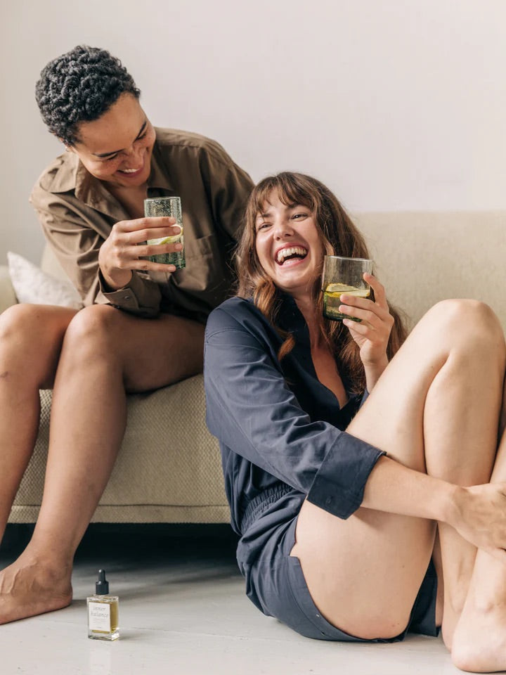 Zwei Frauen genießen INNER BALANCE Detox-Drink in entspannter Atmosphäre, lachend auf dem Boden sitzend